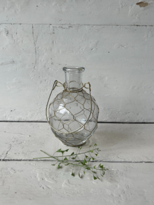 vaso trasparente di forma arrotondata che si restringe verso l'apertura, che è stretta e cilindrica, avvolto da un supporto in filo metallico dorato che forma una rete decorativa intorno al vetro.Dotato di un manico che permette di appendere il vaso.